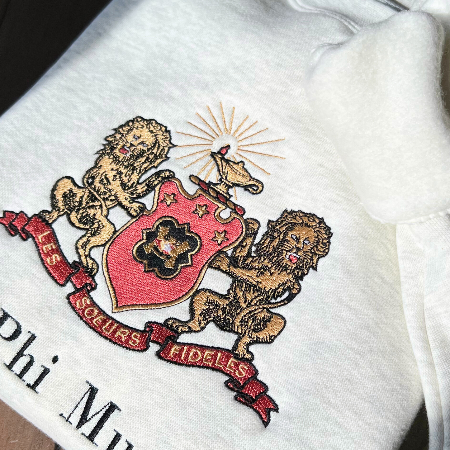 Detailed view of Phi Mu's crest embroidered on a light gray sweatshirt, featuring a shield guarded by gold lions. The shield includes the phi mu badge and under it is Phi Mu's open motto 'Les Soeurs Fideles' which translates to 'the Faithful Sisters'. Above the shield is the crest, the lighted lamp of knowledge in gold. Below the sorority name is embroidered in black.