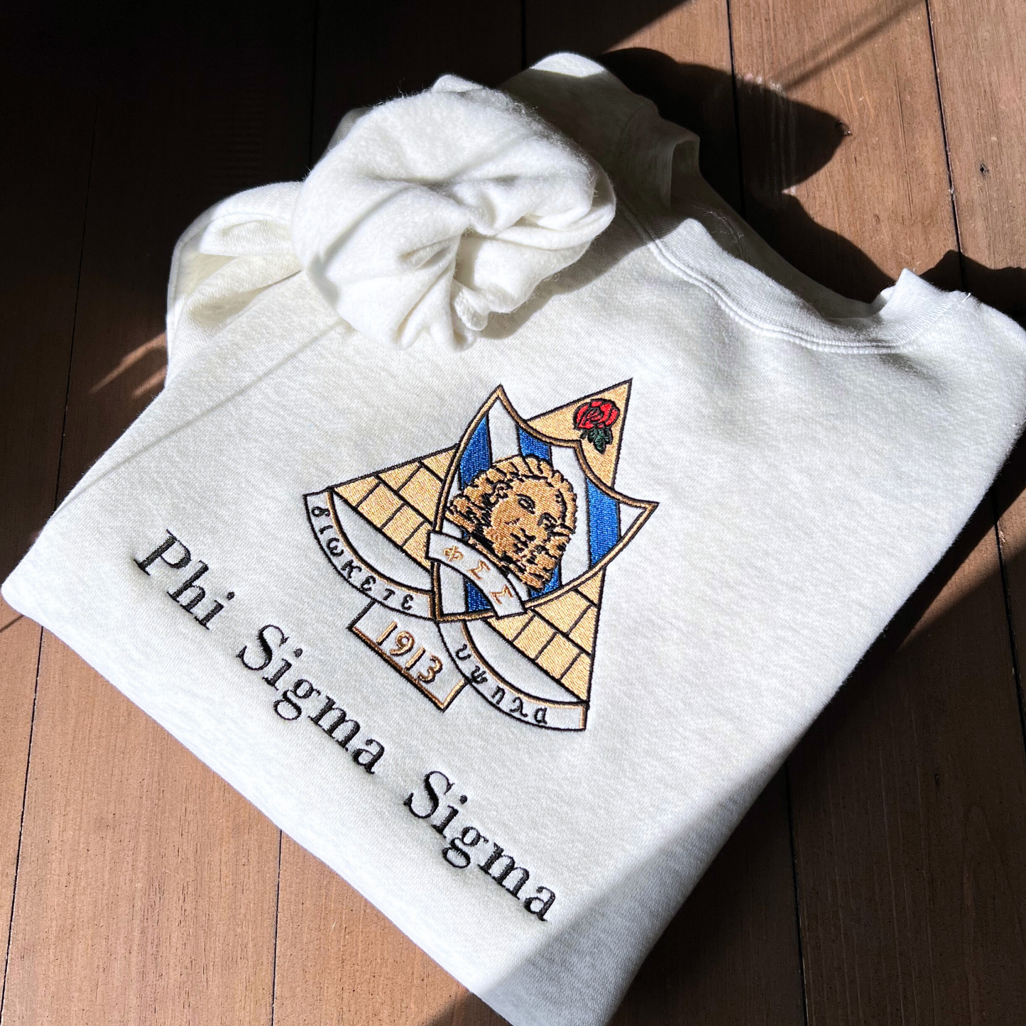 Phi Sigma Sigma's crest embroidered on a light grey sweatshirt, featuring a sphinx head on a bold base with sapphire eyes and the greek letters. It includes the sorority motto "Diokete Hupsula" which translates to "aim high". Below the sorority name is embroidered in black.