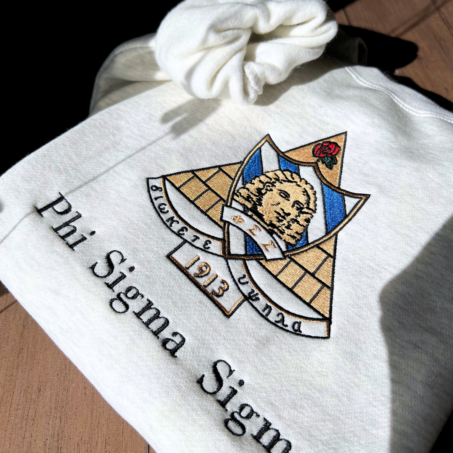Detailed view of Phi Sigma Sigma's crest embroidered on a light grey sweatshirt, featuring a sphinx head on a bold base with sapphire eyes and the greek letters. It includes the sorority motto "Diokete Hupsula" which translates to "aim high". Below the sorority name is embroidered in black.