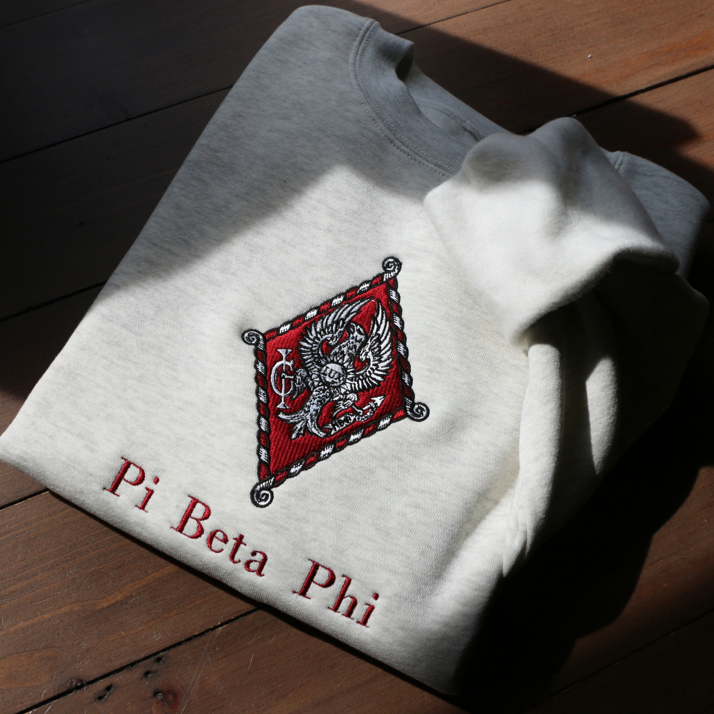 Pi Beta Phi's crest embroidered on a light grey sweatshirt. Featuring an eagle, the seal of Monmouth College, the blazing sun with the latin word lux within, and the Brownlee family crest. Below, the sorority name is embroidered.