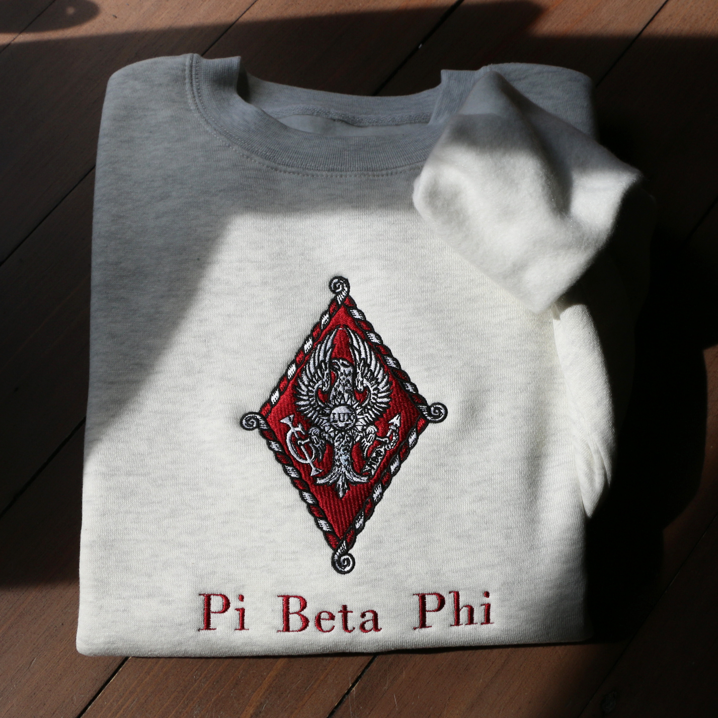Pi Beta Phi's crest embroidered on a light grey sweatshirt. Featuring an eagle, the seal of Monmouth College, the blazing sun with the latin word lux within, and the Brownlee family crest. Below, the sorority name is embroidered.