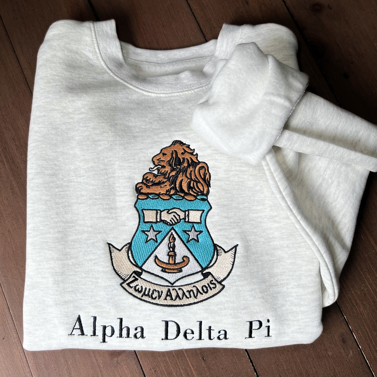 Gray crewneck sweatshirt featuring the Alpha Delta Pi sorority crest embroidered in the center. The crest includes a brown lion atop a blue shield with white stars. Below, the sorority name is embroidered in black.