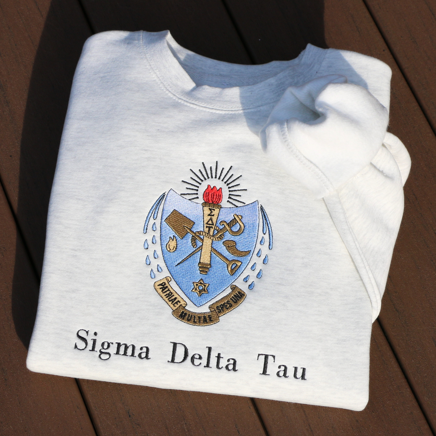 Sigma Delta Tau's crest embroidered on a light grey sweatshirt. Featuring a light blue and gold shield with a red flame and torch, flanked by seven drops to either side and the motto "Patriae Multae spes Una" which translates to "One Hope of Many People" in a ribbon along the bottom. Below the sorority name is embroidered in black.