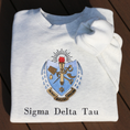 Load image into Gallery viewer, Sigma Delta Tau's crest embroidered on a light grey sweatshirt. Featuring a light blue and gold shield with a red flame and torch, flanked by seven drops to either side and the motto "Patriae Multae spes Una" which translates to "One Hope of Many People" in a ribbon along the bottom. Below the sorority name is embroidered in black.