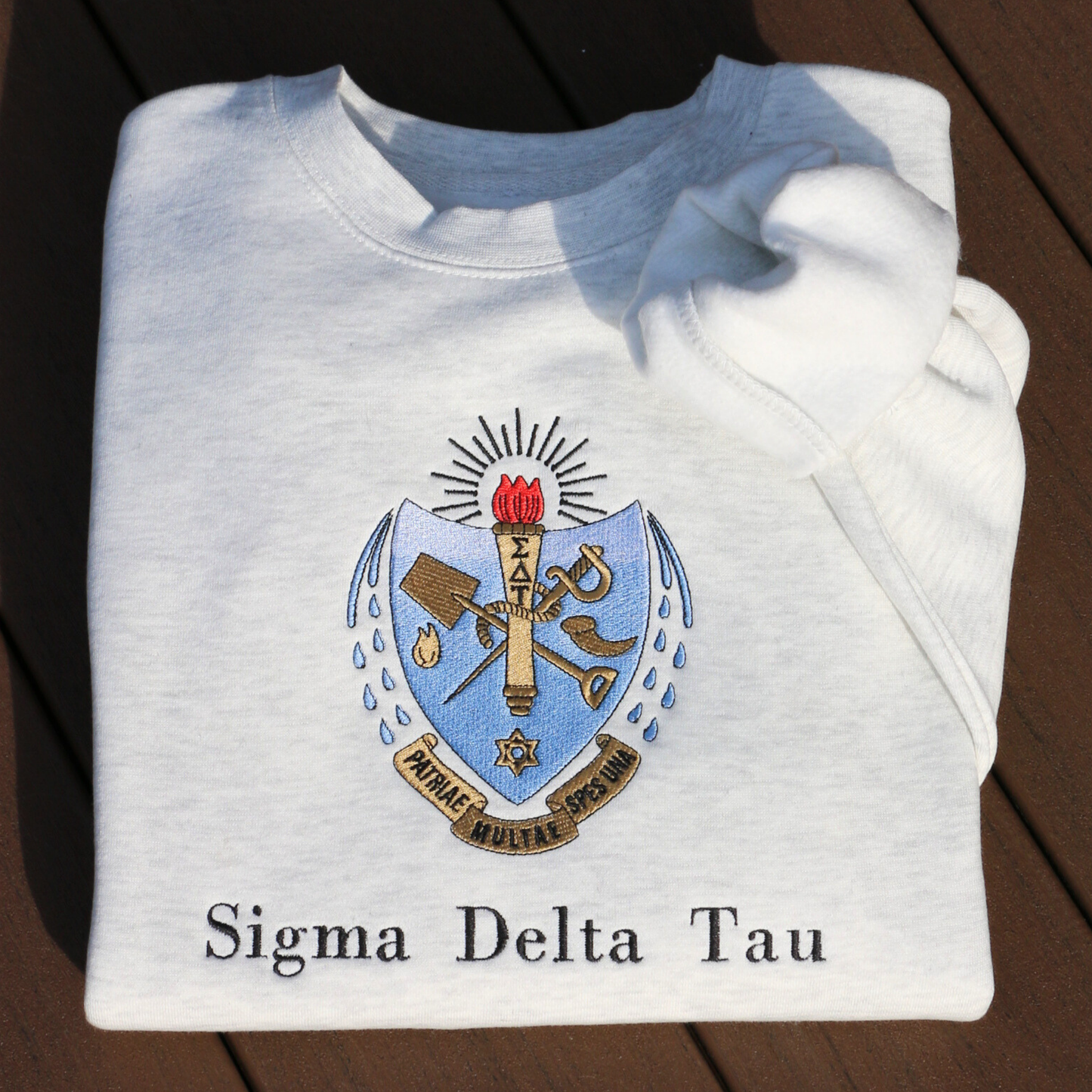 Sigma Delta Tau's crest embroidered on a light grey sweatshirt. Featuring a light blue and gold shield with a red flame and torch, flanked by seven drops to either side and the motto "Patriae Multae spes Una" which translates to "One Hope of Many People" in a ribbon along the bottom. Below the sorority name is embroidered in black.