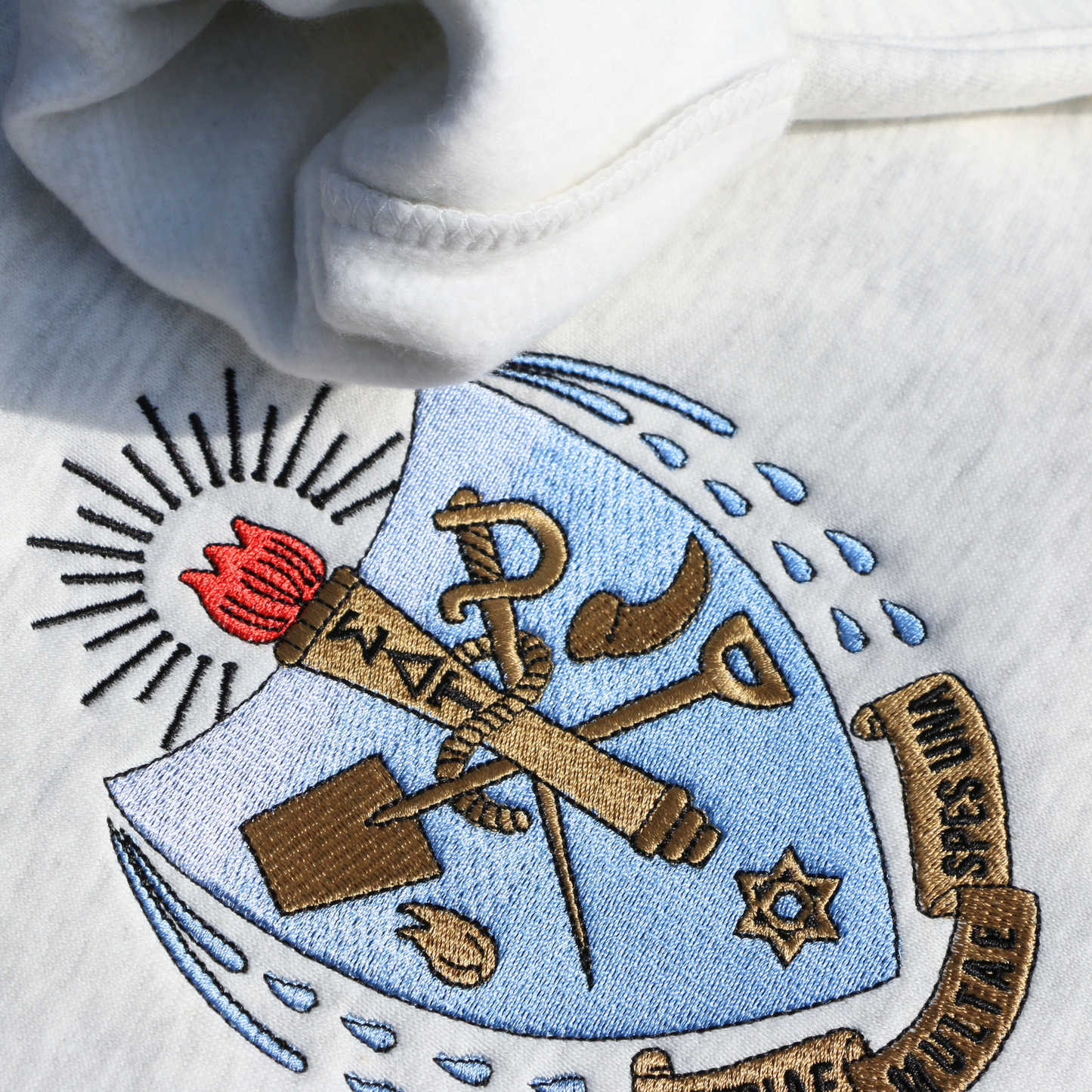 Detailed view of Sigma Delta Tau's crest embroidered on a light grey sweatshirt. Featuring a light blue and gold shield with a red flame and torch, flanked by seven drops to either side and the motto "Patriae Multae spes Una" which translates to "One Hope of Many People" in a ribbon along the bottom. Below the sorority name is embroidered in black.