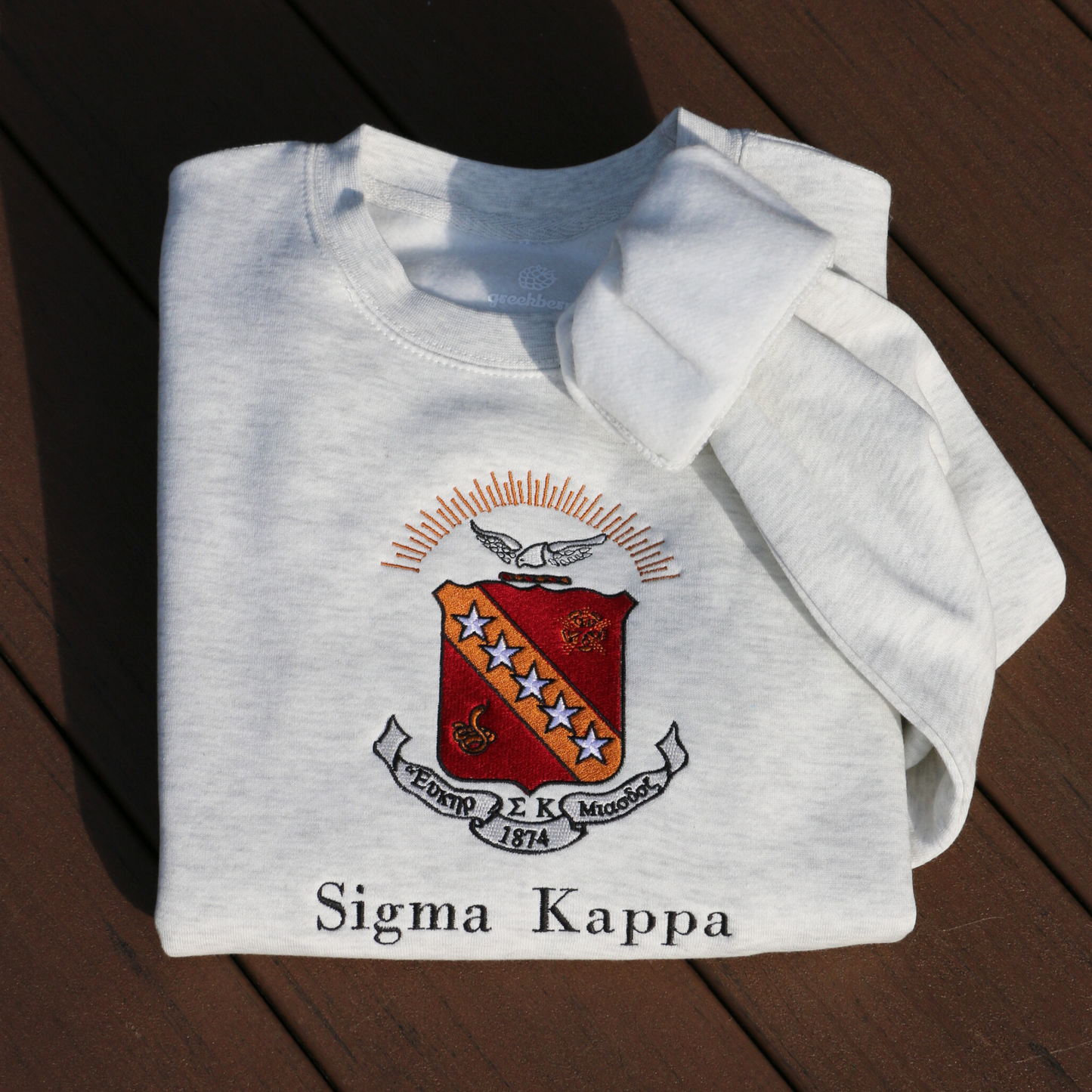 Sigma Kappa's crest embroidered on a light grey sweatshirt. Featuring a maroon shield with a diagonal gold bar on which are five stars. The lower half has a coiled serpent, and the upper has a maroon and gold wreath. Below, the sorority's name is embroidered in black.