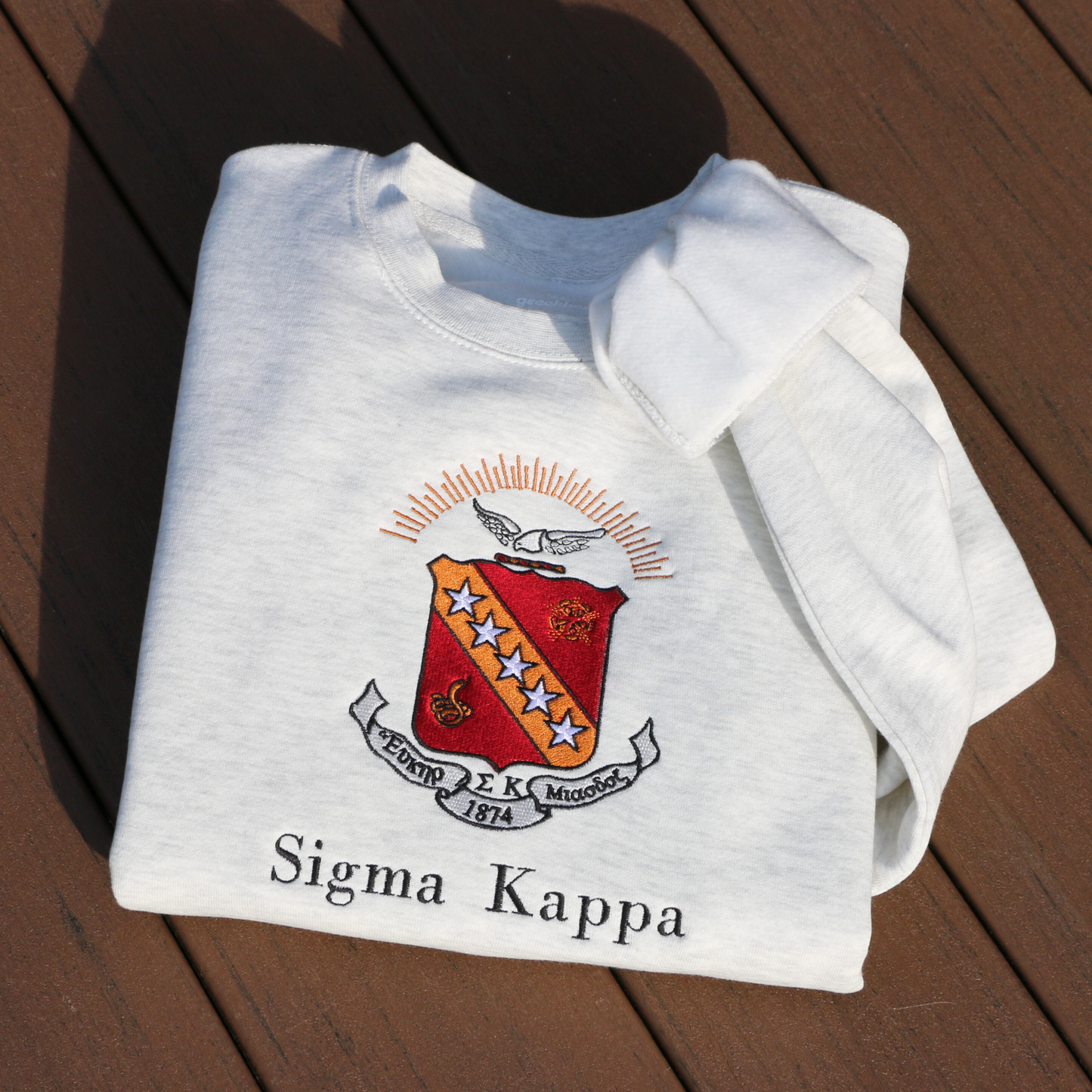 Sigma Kappa's crest embroidered on a light grey sweatshirt. Featuring a maroon shield with a diagonal gold bar on which are five stars. The lower half has a coiled serpent, and the upper has a maroon and gold wreath. Below, the sorority's name is embroidered in black.
