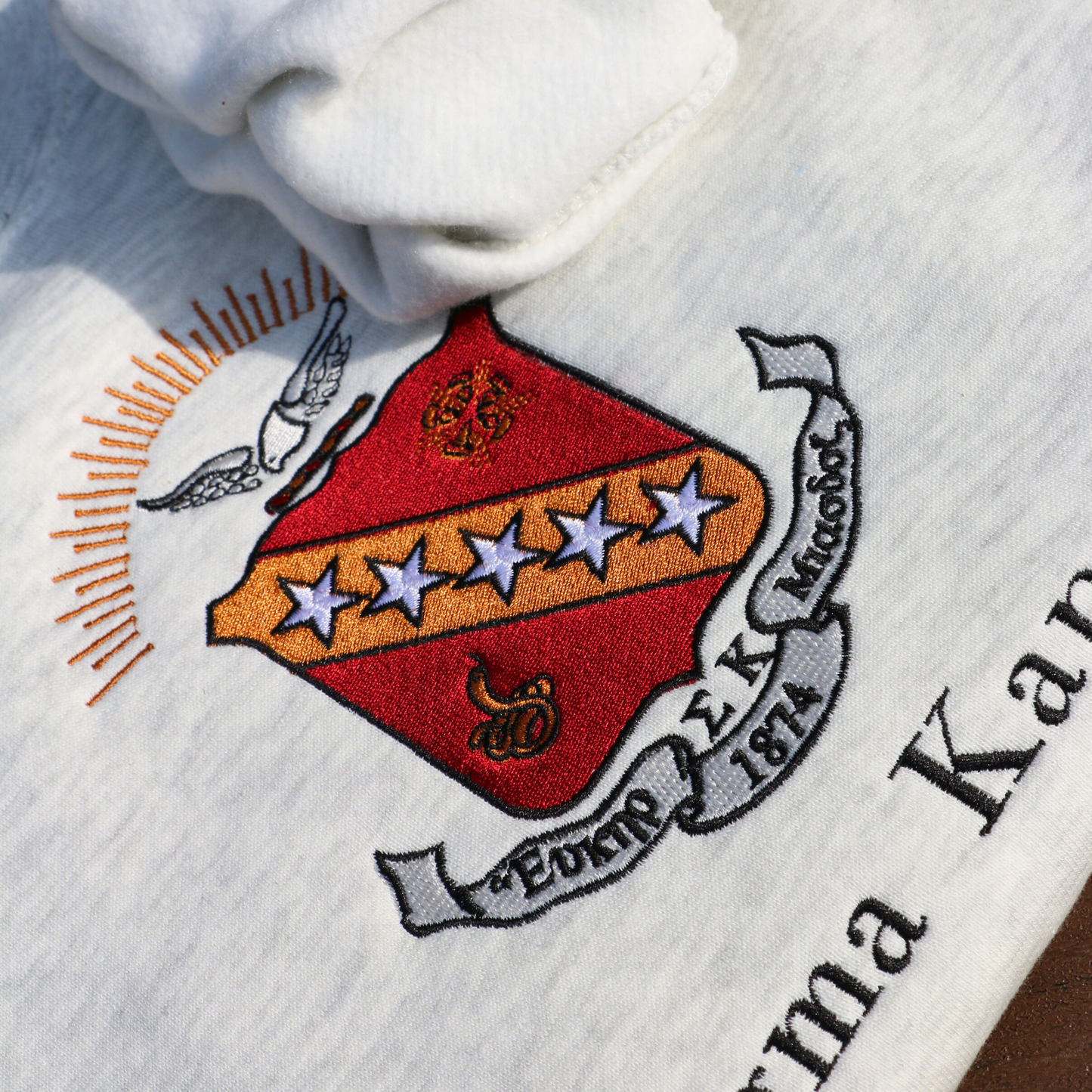 Detailed view of Sigma Kappa's crest embroidered on a light grey sweatshirt. Featuring a maroon shield with a diagonal gold bar on which are five stars. The lower half has a coiled serpent, and the upper has a maroon and gold wreath. Below, the sorority's name is embroidered in black.