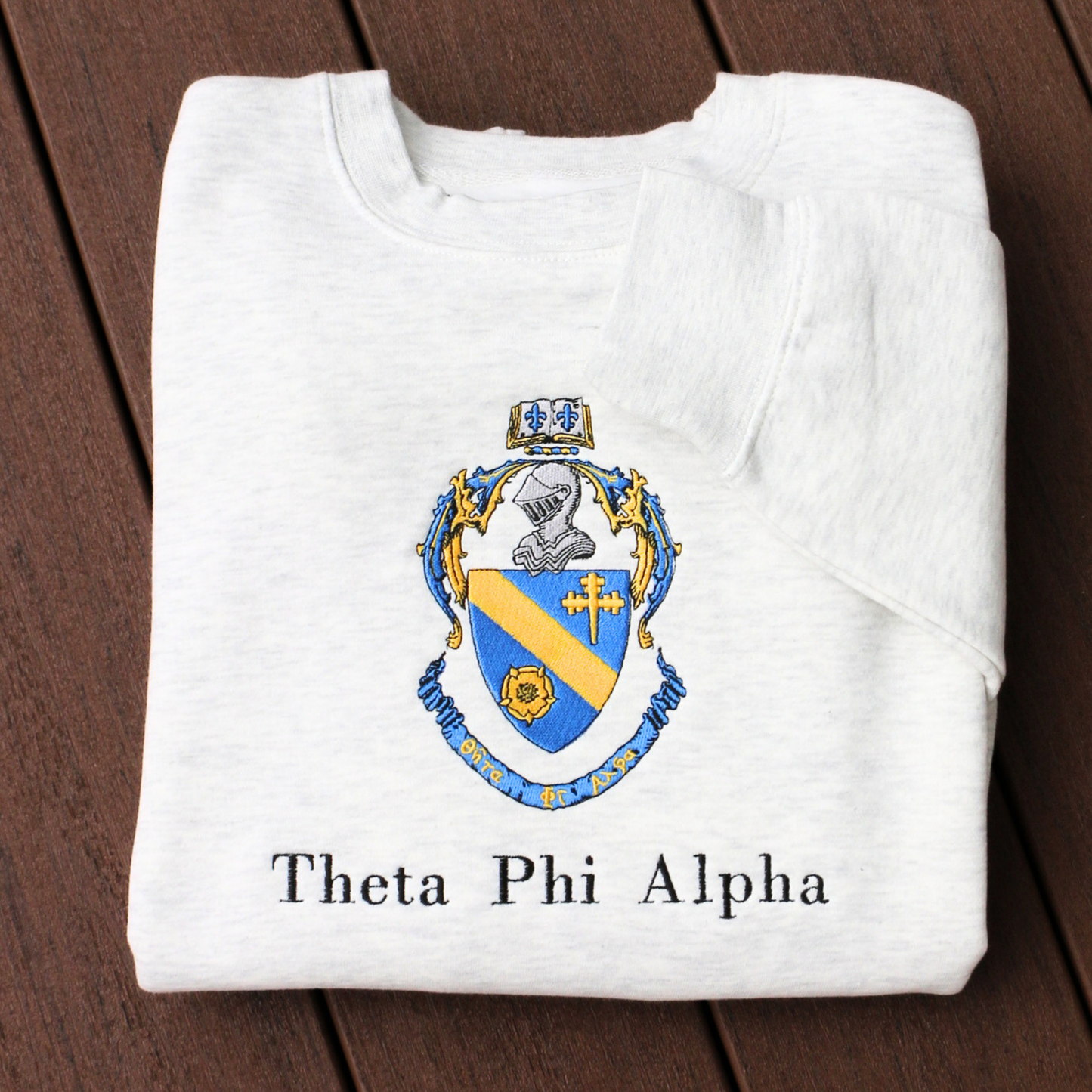 Theta Phi Alpha's crest embroidered on a light grey sweatshirt. Featuring a gold and blue crest, with an open book and head of armor atop it. The book is imprinted with two blue fleur-de-lis. The motto is written on the blue banner at the bottom of the crest. The sorority name is embroidered beneath in black.