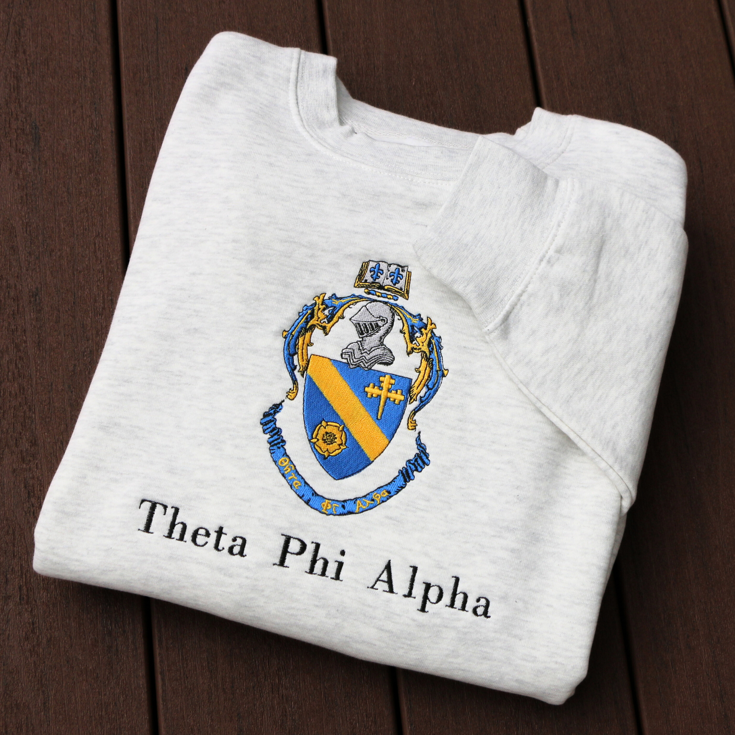 Theta Phi Alpha's crest embroidered on a light grey sweatshirt. Featuring a gold and blue crest, with an open book and head of armor atop it. The book is imprinted with two blue fleur-de-lis. The motto is written on the blue banner at the bottom of the crest. The sorority name is embroidered beneath in black.