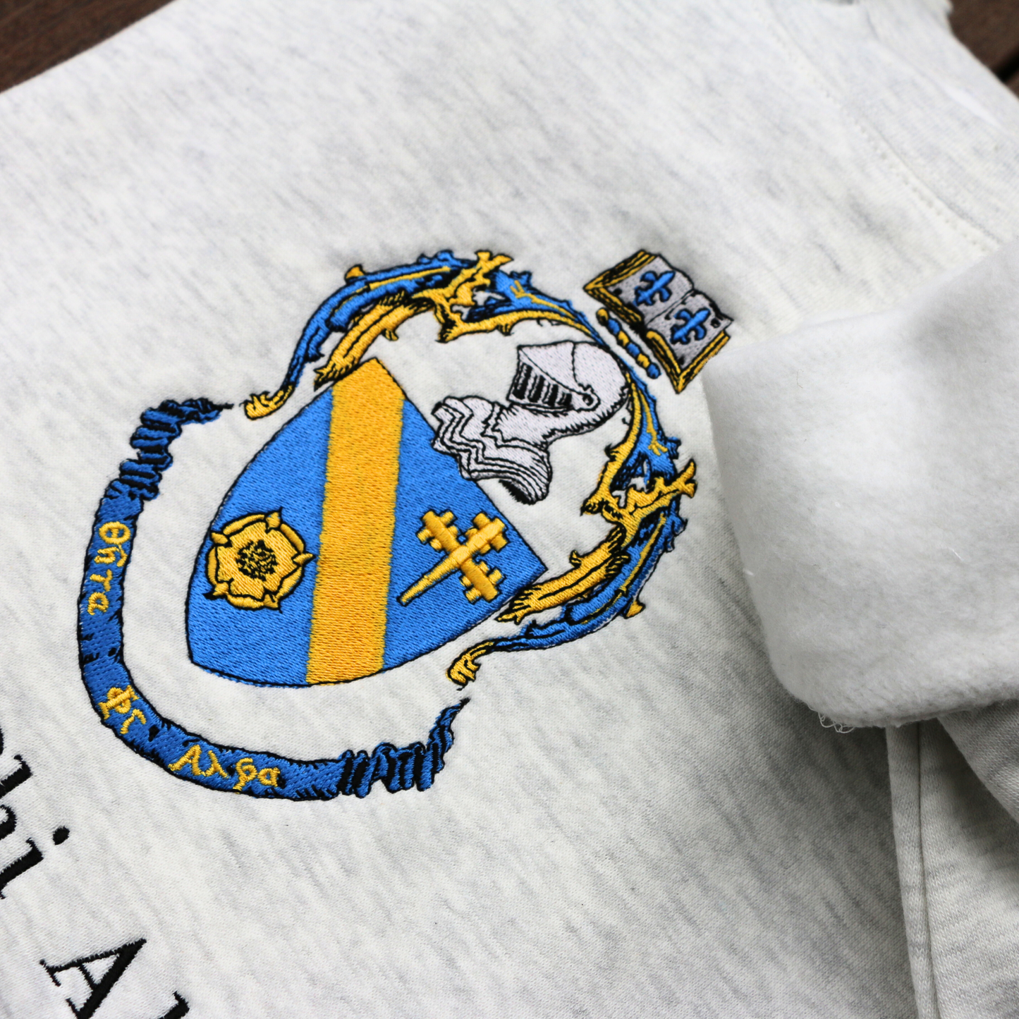 Detailed view of Theta Phi Alpha's crest embroidered on a light grey sweatshirt. Featuring a gold and blue crest, with an open book and head of armor atop it. The book is imprinted with two blue fleur-de-lis. The motto is written on the blue banner at the bottom of the crest. The sorority name is embroidered beneath in black.