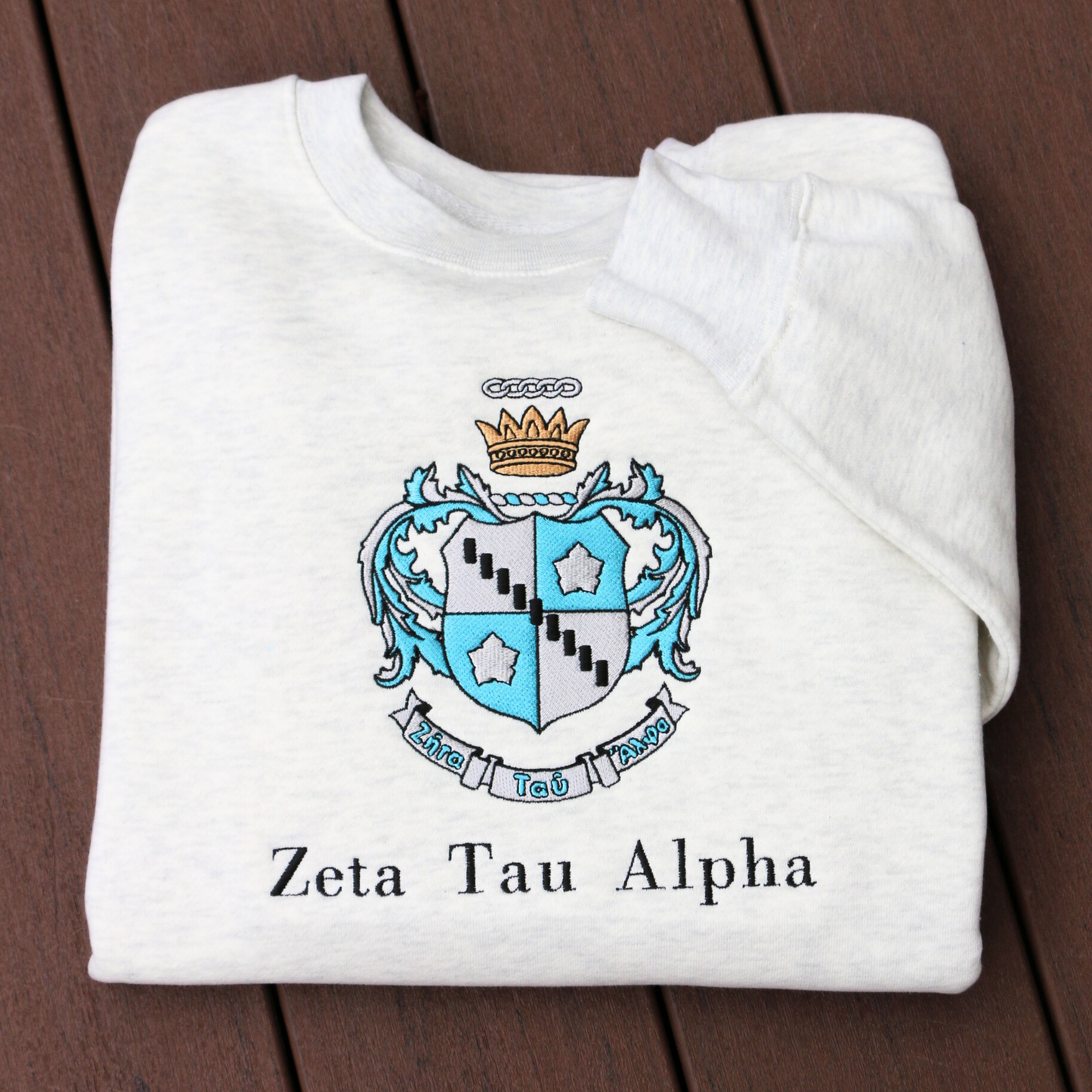 Zeta Tau Alpha's crest embroidered on a light grey sweatshirt. Featuring a silver and blue shield with a five pointed crown. The sorority name 'Zeta Tau Alpha' is embroidered in black beneath.