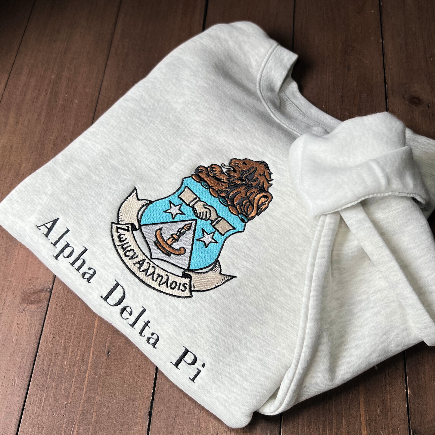 Gray crewneck sweatshirt featuring the Alpha Delta Pi sorority crest embroidered in the center. The crest includes a brown lion atop a blue shield with white stars. Below, the sorority name is embroidered in black.