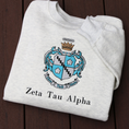 Load image into Gallery viewer, Zeta Tau Alpha's crest embroidered on a light grey sweatshirt. Featuring a silver and blue shield with a five pointed crown. The sorority name 'Zeta Tau Alpha' is embroidered in black beneath.