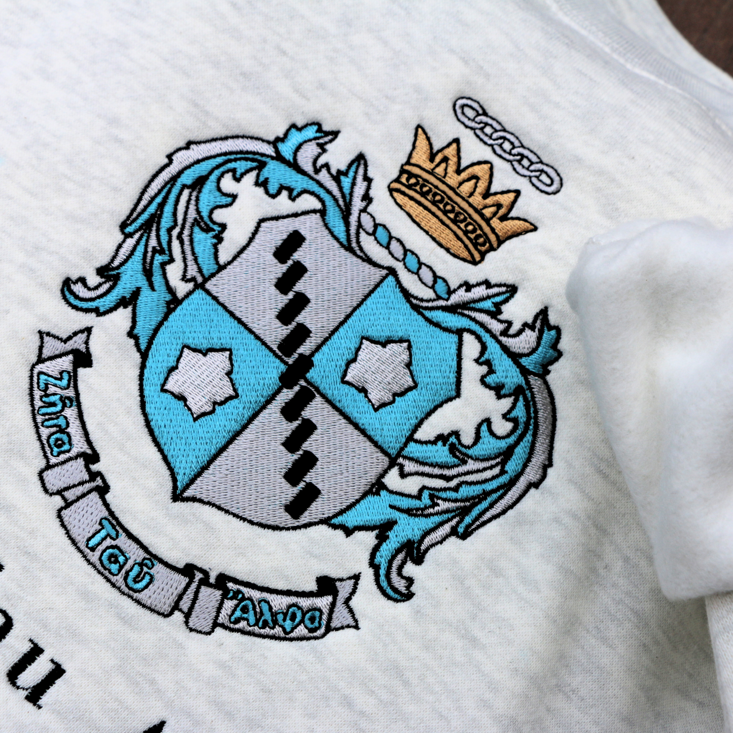 Detailed view of Zeta Tau Alpha's crest embroidered on a light grey sweatshirt. Featuring a silver and blue shield with a five pointed crown. The sorority name 'Zeta Tau Alpha' is embroidered in black beneath.