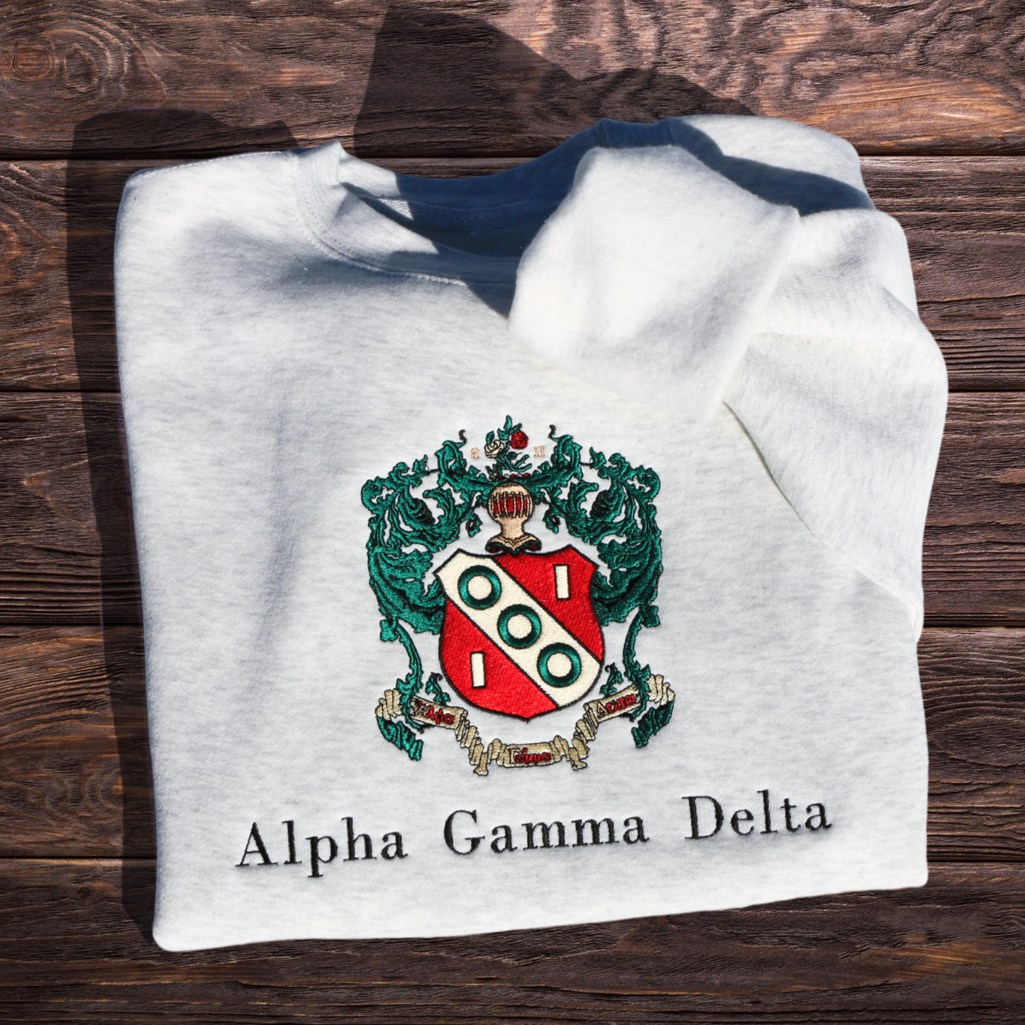 Gray crewneck sweatshirt laid flat on a wooden surface, featuring a detailed embroidery of the Alpha Gamma Delta sorority crest in vibrant colors. The crest includes a red shield surrounded by green leaves. Below the crest, 'Alpha Gamma Delta' is embroidered in black script