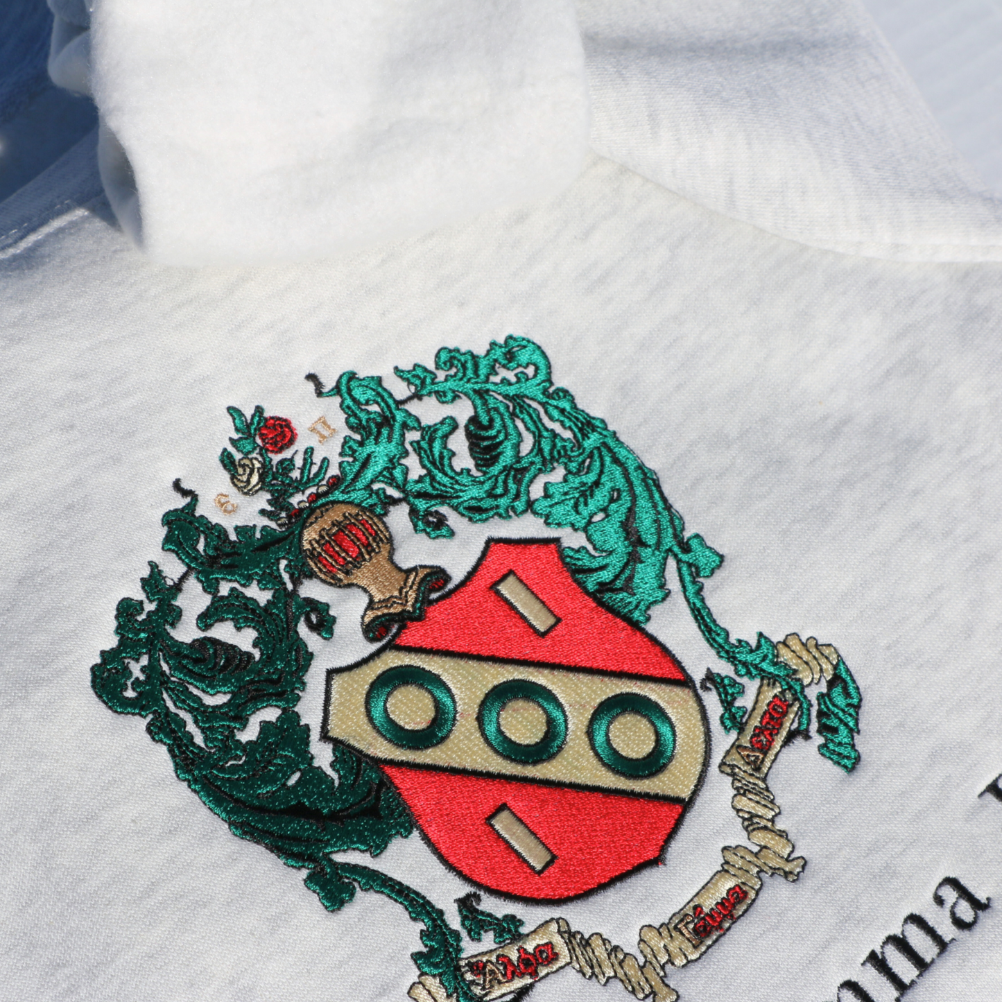 Close-up view of the detailed embroidery on a gray crewneck sweatshirt featuring the Alpha Gamma Delta sorority crest. The crest showcases a red shield surrounded by intricate green leaves.