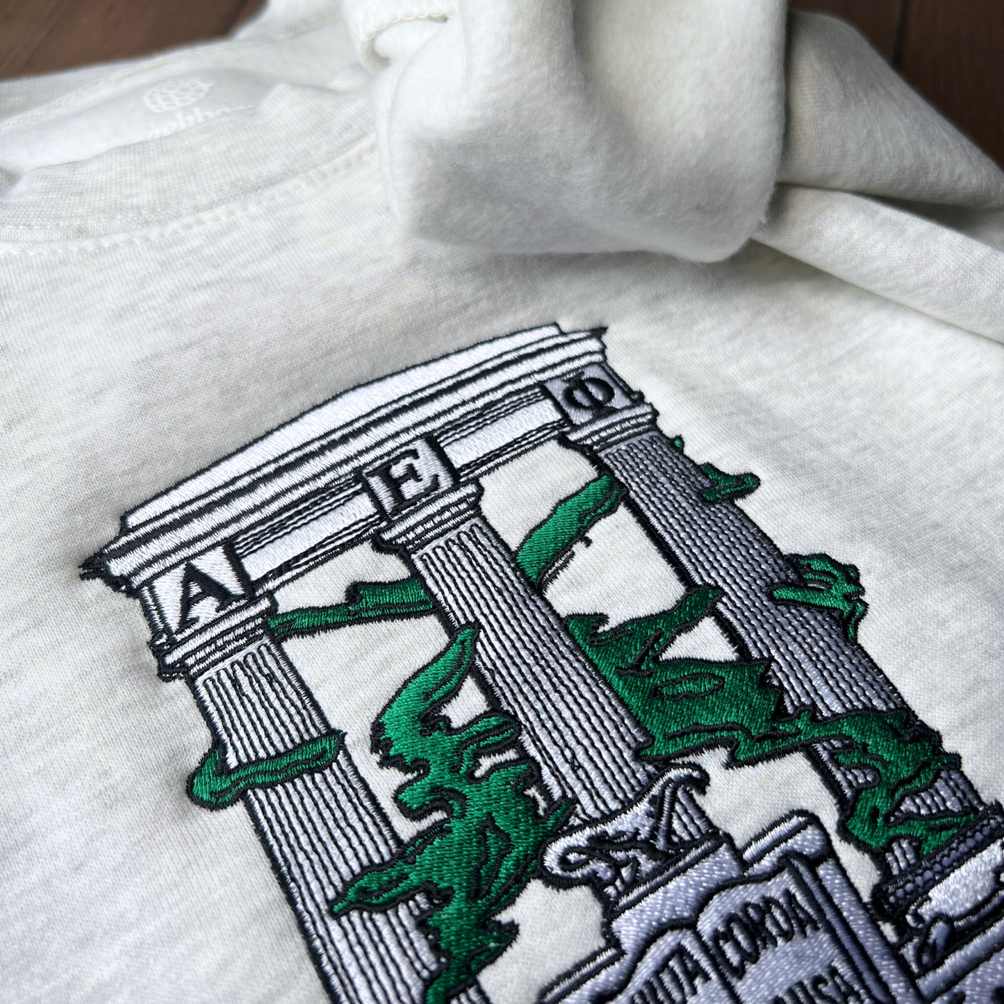 A detailed view of a folded light gray sweatshirt with an embroidered design of Alpha Epsilon Phi's emblem featuring white columns and green foliage. The columns support a structure with the Greek letters 'ΑΕΦ'. Below, the motto 'Multa Corda, Una Causa' is prominently displayed.