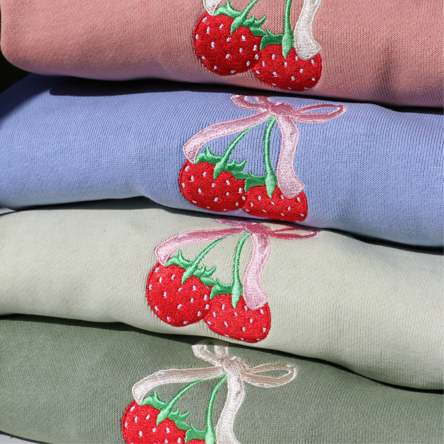 Stack of folded sweatshirts in peach, glacier (light blue), kiwi (light green), and eucalyptus (green), each embroidered with red strawberries tied together with a pink or cream bow.