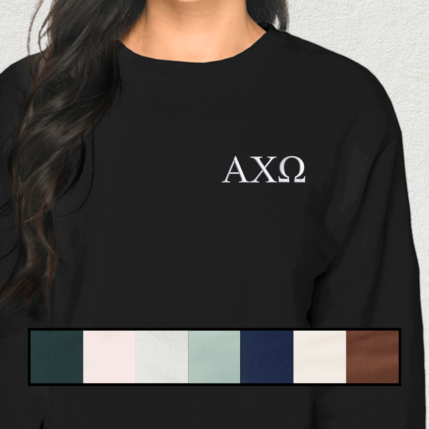 A young woman wearing a black sweatshirt with her sorority letters embroidered in white on the chest. The sweatshirt has a classic crew neck and long sleeves, displayed on a neutral textured background.