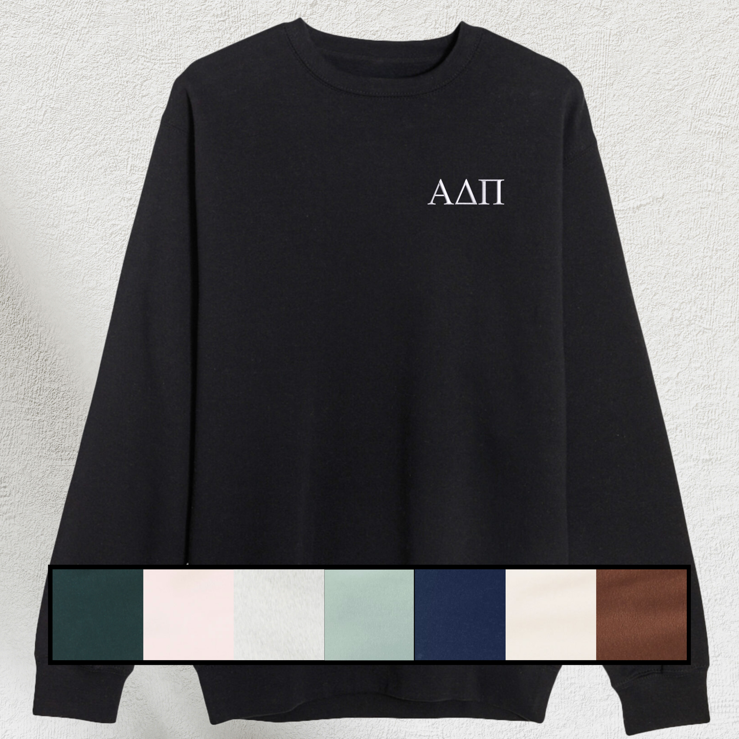 A black sweatshirt laid flat, showing sorority letters embroidered in white on the chest. Below, a swatch palette of available sweatshirt colors including forest, pale pink, seafoam, navy, chestnut, grey, cream, and black is visible.