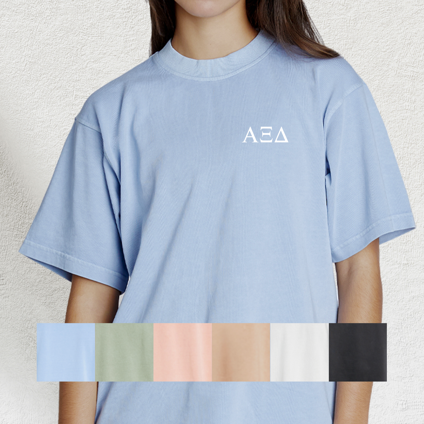 A woman wearing a light blue t-shirt with the Greek letters of her sorority on it in white on the front. The image includes a color swatch below with shades light blue, light green, peach, brown, grey, and black.