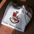 Load image into Gallery viewer, Light gray crewneck sweatshirt laid flat on a wooden surface, showcasing the Alpha Sigma Alpha sorority crest embroidered in the center. The crest displays a red shield with a golden phoenix, topped by a crown, and surrounded by a scroll bearing the sorority's Greek motto and name.
