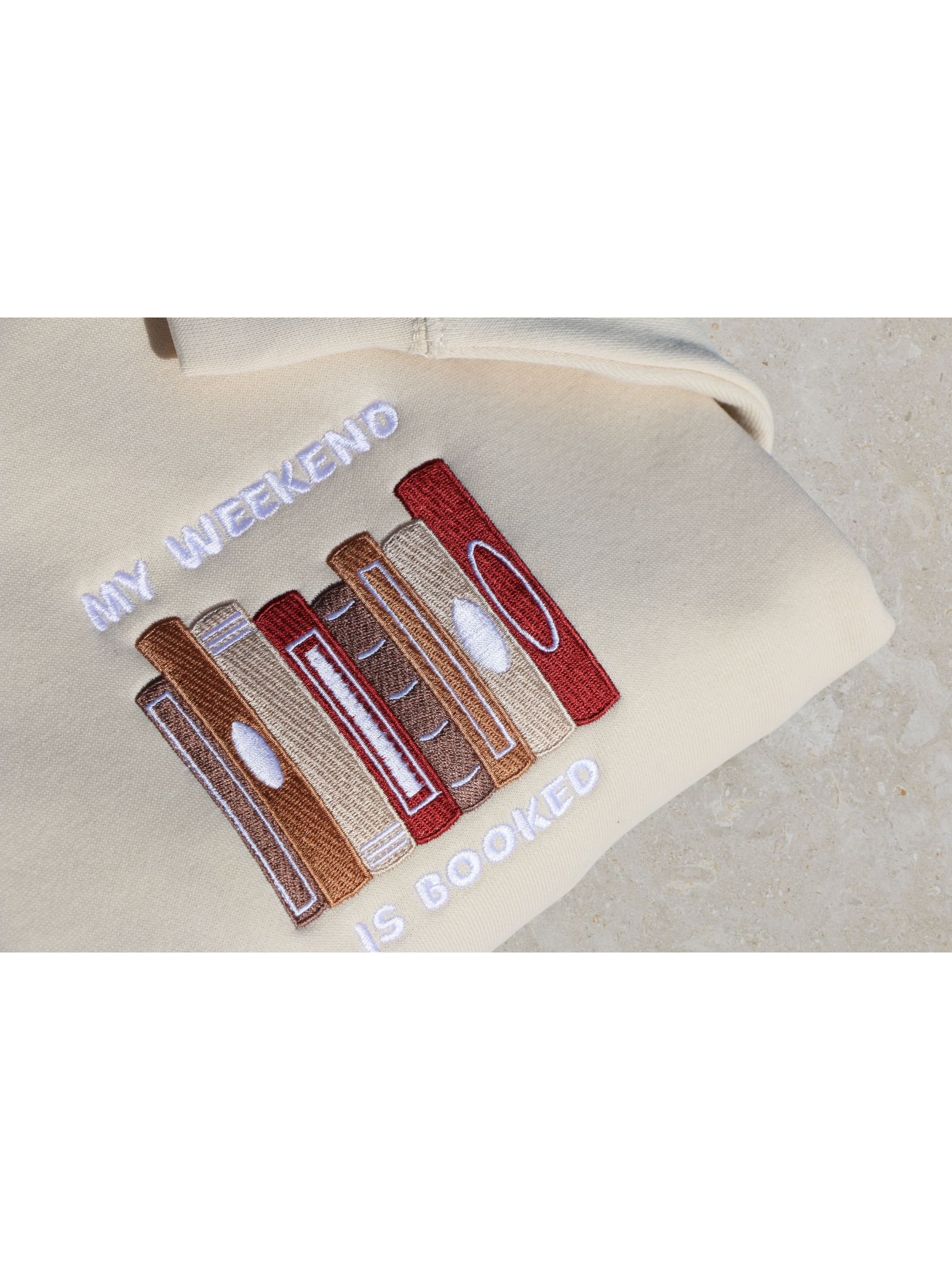 Close-up view of a beige sweatshirt embroidered with a design of multiple book spines in various brown, red, and gold shades, and the phrase 'My Weekend is Booked'. Set against a neutral background.