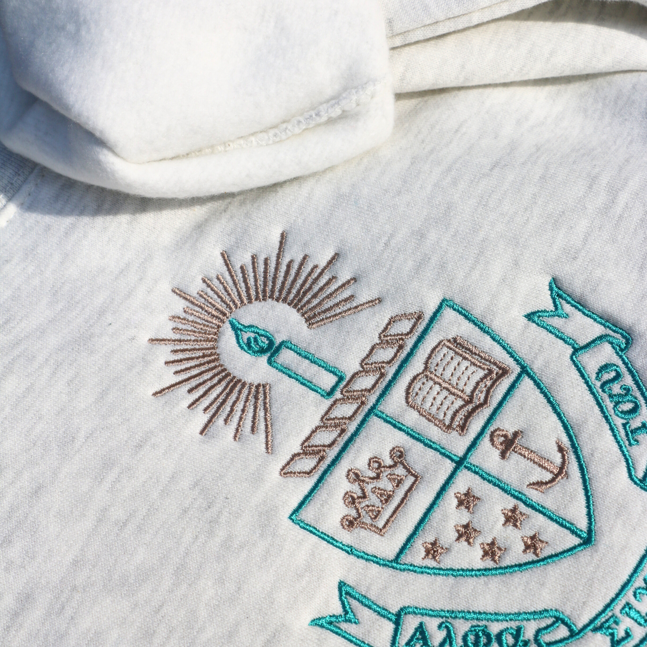 Detailed view of a light gray crewneck sweatshirt laid flat on a dark wood surface, featuring the embroidered crest of Alpha Sigma Tau. The crest includes a shield divided into four, showcasing a book, a crown, an anchor, and stars in teal and gold, with a candle and a golden light above. Below, the sorority name 'Alpha Sigma Tau' is embroidered below in gold.