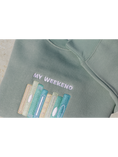 Load image into Gallery viewer, Close-up view of a mint green sweatshirt embroidered with a design of multiple book spines in various blue, green, and yellow shades, and the phrase 'My Weekend is Booked'. Set against a neutral background.
