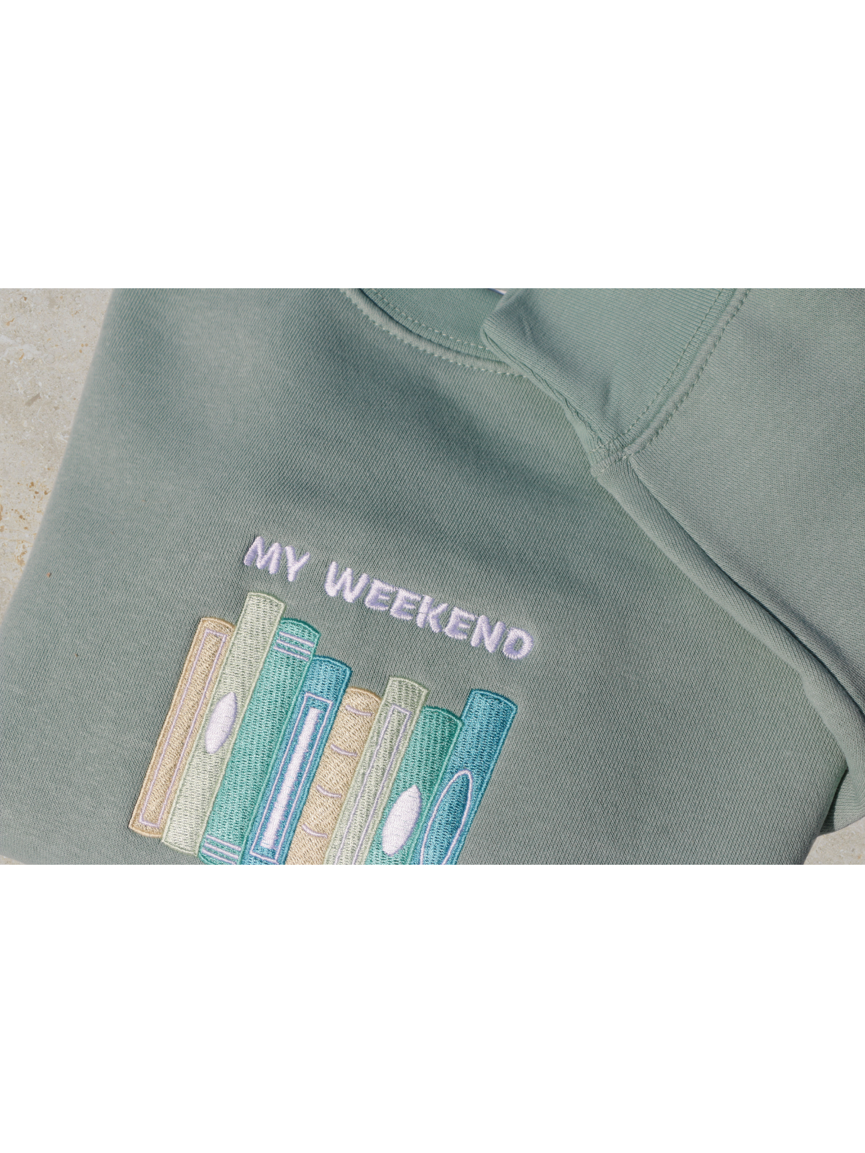 Close-up view of a mint green sweatshirt embroidered with a design of multiple book spines in various blue, green, and yellow shades, and the phrase 'My Weekend is Booked'. Set against a neutral background.