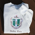Load image into Gallery viewer, Delta Zeta's crest embroidered on a light gray sweatshirt, featuring a green shield with a roman lamp and scroll. Below, the sorority's name is embroidered in brown.
