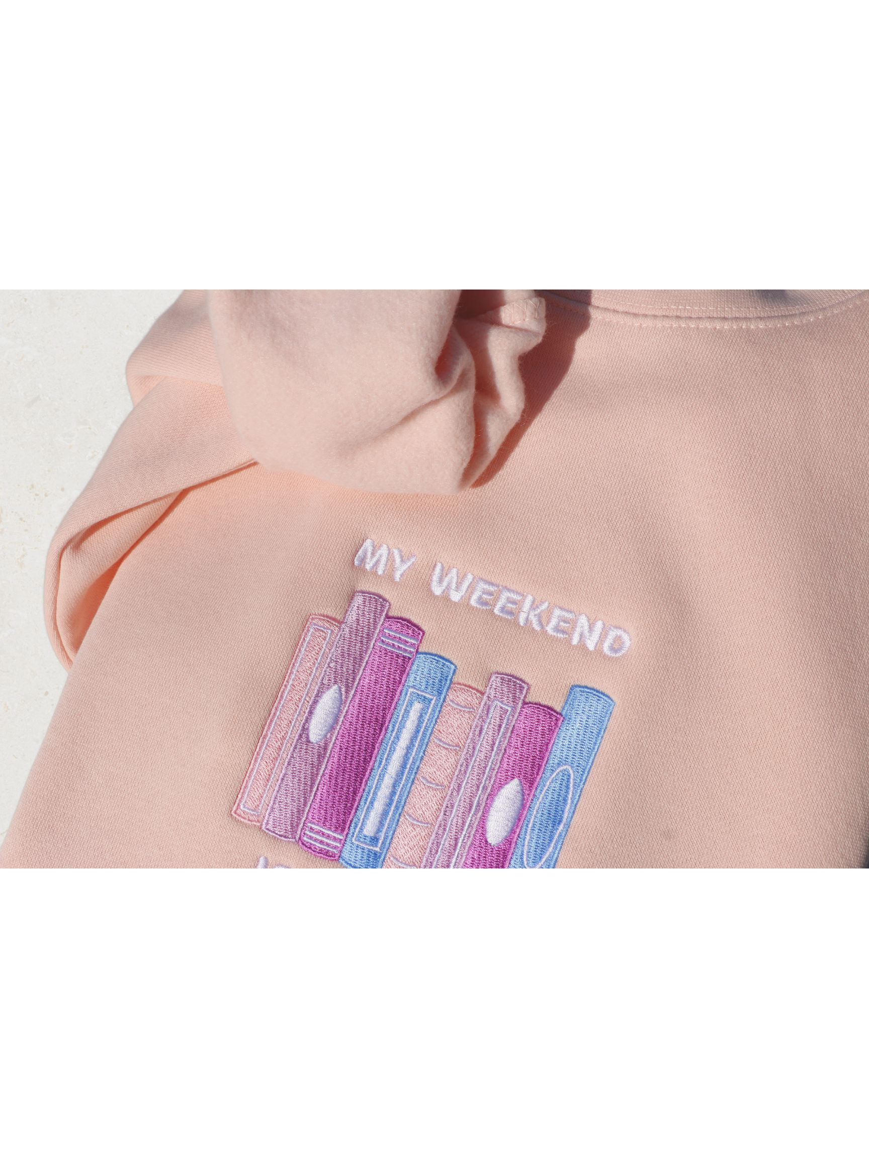 Close-up view of a pink sweatshirt embroidered with a design of multiple book spines in various pink, purple, and blue shades, and the phrase 'My Weekend is Booked'. Set against a neutral background.