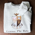 Load image into Gallery viewer, Gamma Phi Beta's crest embroidered on a light gray sweatshirt, featuring a brown shield with a golden crescent moon and pink flowers, and an open book, topped by a golden lamp, with 'Gamma Phi Beta' written below in elegant script.
