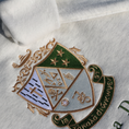 Load image into Gallery viewer, Detailed view of Kappa Delta's crest embroidered on a light gray sweatshirt, featuring a green and gold shield with stars, a skull and crossbones, a cross, the sorority's greek letters, a lamp, and a scroll. a white rose and stars, underlined by a scroll with 'Τa Kala Diokomen' written on it. Below, the sorority name is embroidered in dark green.
