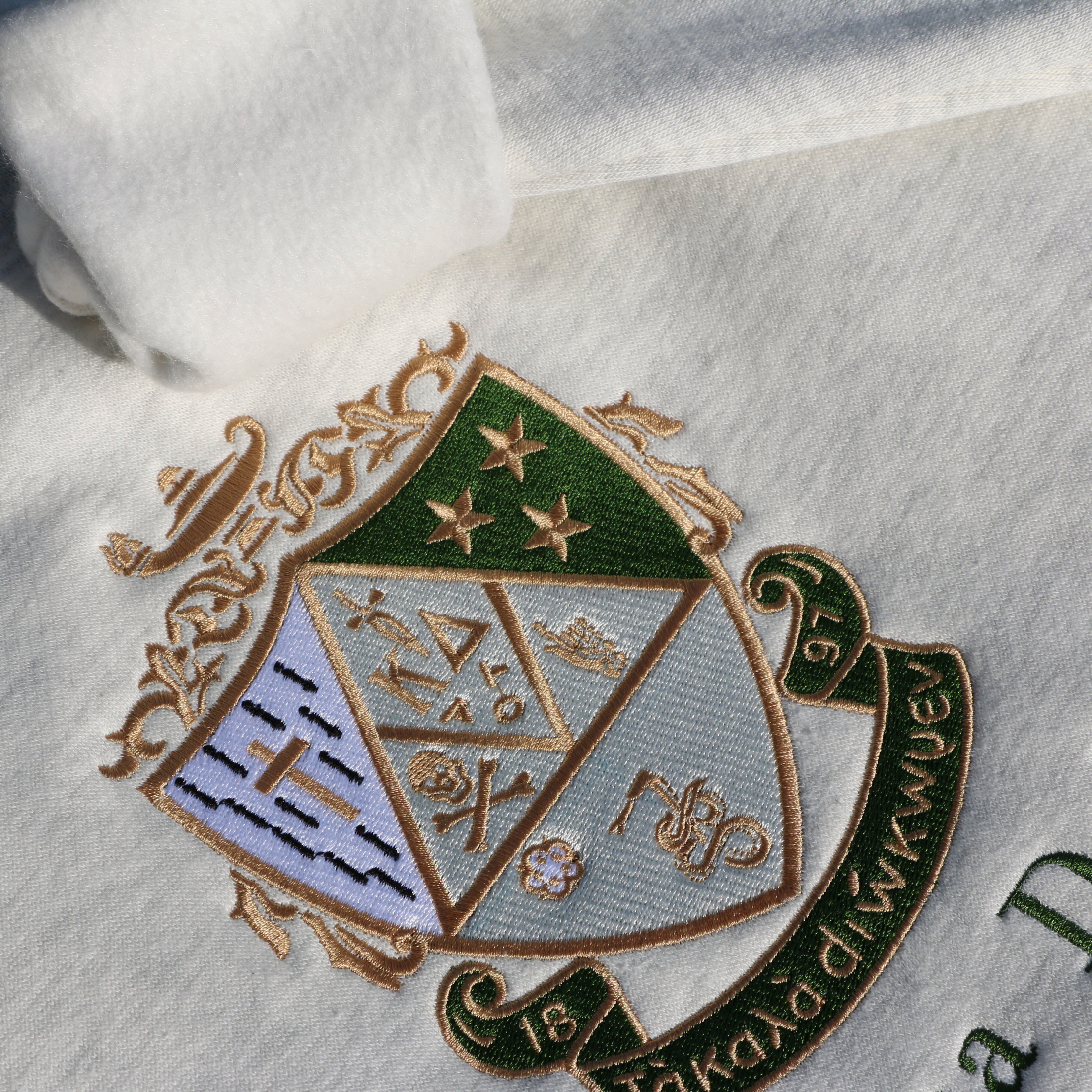 Detailed view of Kappa Delta's crest embroidered on a light gray sweatshirt, featuring a green and gold shield with stars, a skull and crossbones, a cross, the sorority's greek letters, a lamp, and a scroll. a white rose and stars, underlined by a scroll with 'Τa Kala Diokomen' written on it. Below, the sorority name is embroidered in dark green.