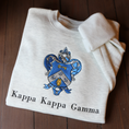Load image into Gallery viewer, Kappa Kappa Gamma's crest embroidered on a light gray sweatshirt, featuring a blue shield with a golden key and fleur-de-lis, with the sorority name 'Kappa Kappa Gamma' in black lettering beneath.
