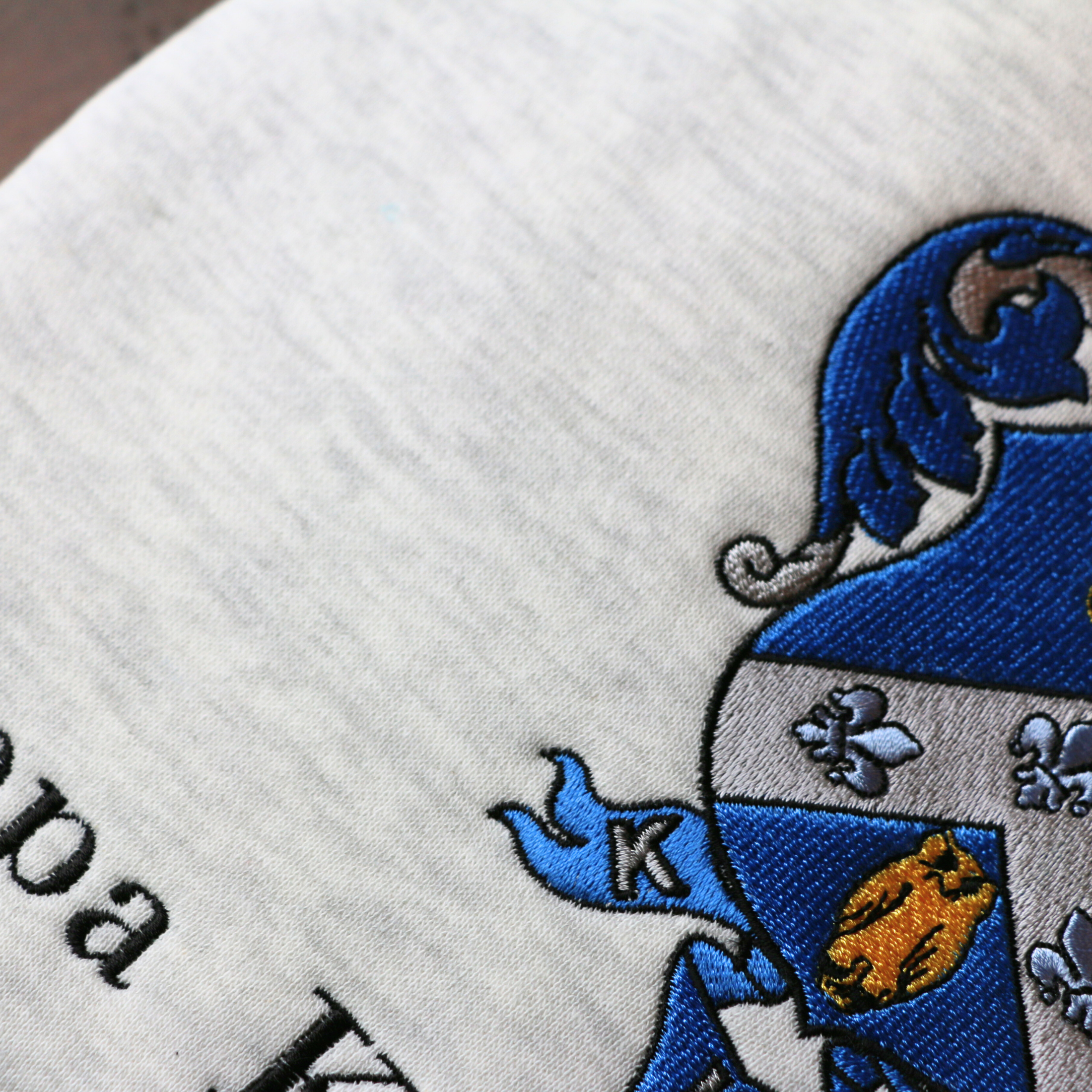 Detailed view of Kappa Kappa Gamma's crest embroidered on a light gray sweatshirt, featuring a blue shield with a golden key and fleur-de-lis, with the sorority name 'Kappa Kappa Gamma' in black lettering beneath.