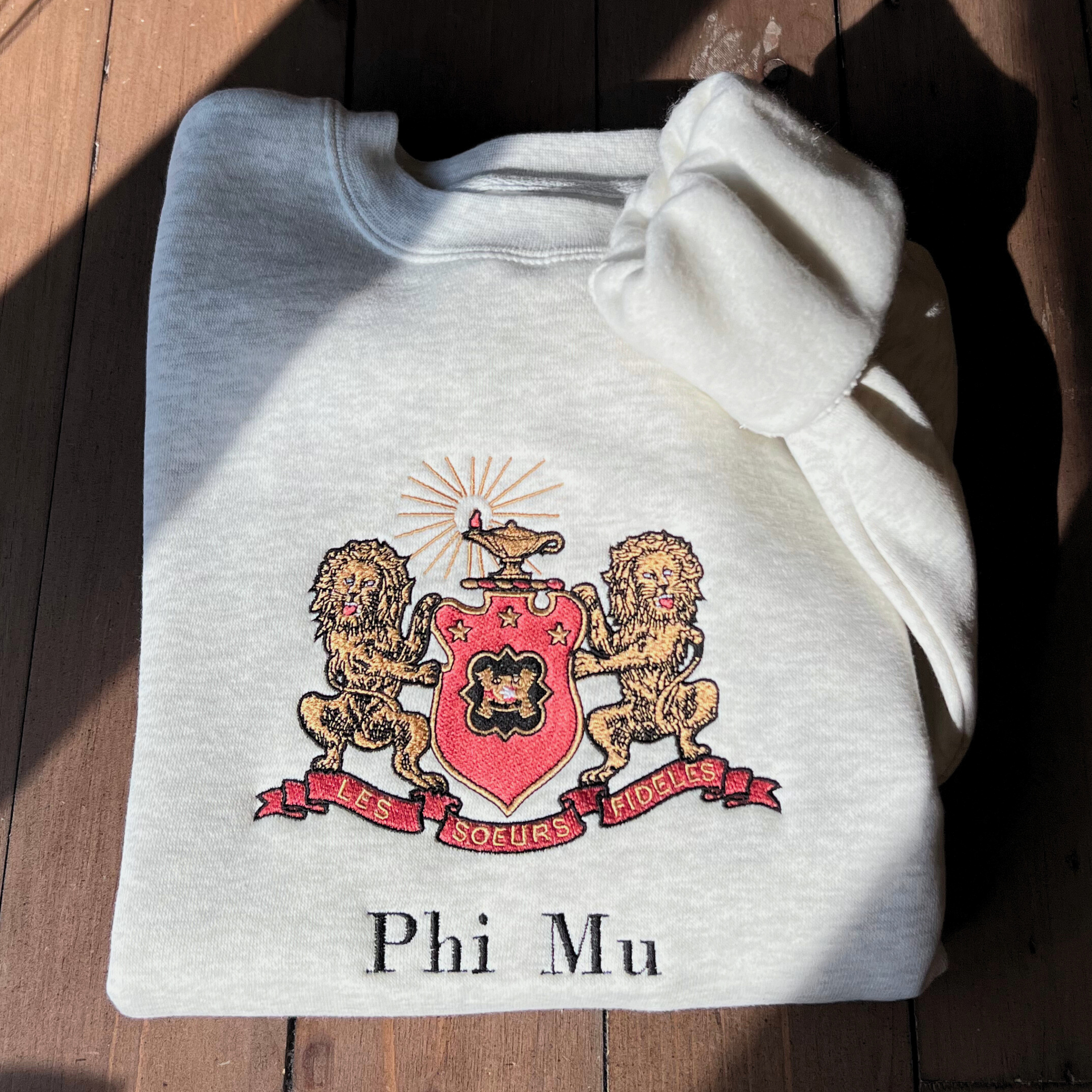 Phi Mu's crest embroidered on a light gray sweatshirt, featuring a shield guarded by gold lions. The shield includes the phi mu badge and under it is Phi Mu's open motto 'Les Soeurs Fideles' which translates to 'the Faithful Sisters'. Above the shield is the crest, the lighted lamp of knowledge in gold. Below the sorority name is embroidered in black.