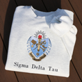 Load image into Gallery viewer, Sigma Delta Tau's crest embroidered on a light grey sweatshirt. Featuring a light blue and gold shield with a red flame and torch, flanked by seven drops to either side and the motto "Patriae Multae spes Una" which translates to "One Hope of Many People" in a ribbon along the bottom. Below the sorority name is embroidered in black.
