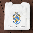 Load image into Gallery viewer, Theta Phi Alpha's crest embroidered on a light grey sweatshirt. Featuring a gold and blue crest, with an open book and head of armor atop it. The book is imprinted with two blue fleur-de-lis. The motto is written  on the blue banner at the bottom of the crest. The sorority name is embroidered beneath in black.
