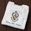 Load image into Gallery viewer, Theta Phi Alpha's crest embroidered on a light grey sweatshirt. Featuring a gold and blue crest, with an open book and head of armor atop it. The book is imprinted with two blue fleur-de-lis. The motto is written  on the blue banner at the bottom of the crest. The sorority name is embroidered beneath in black.
