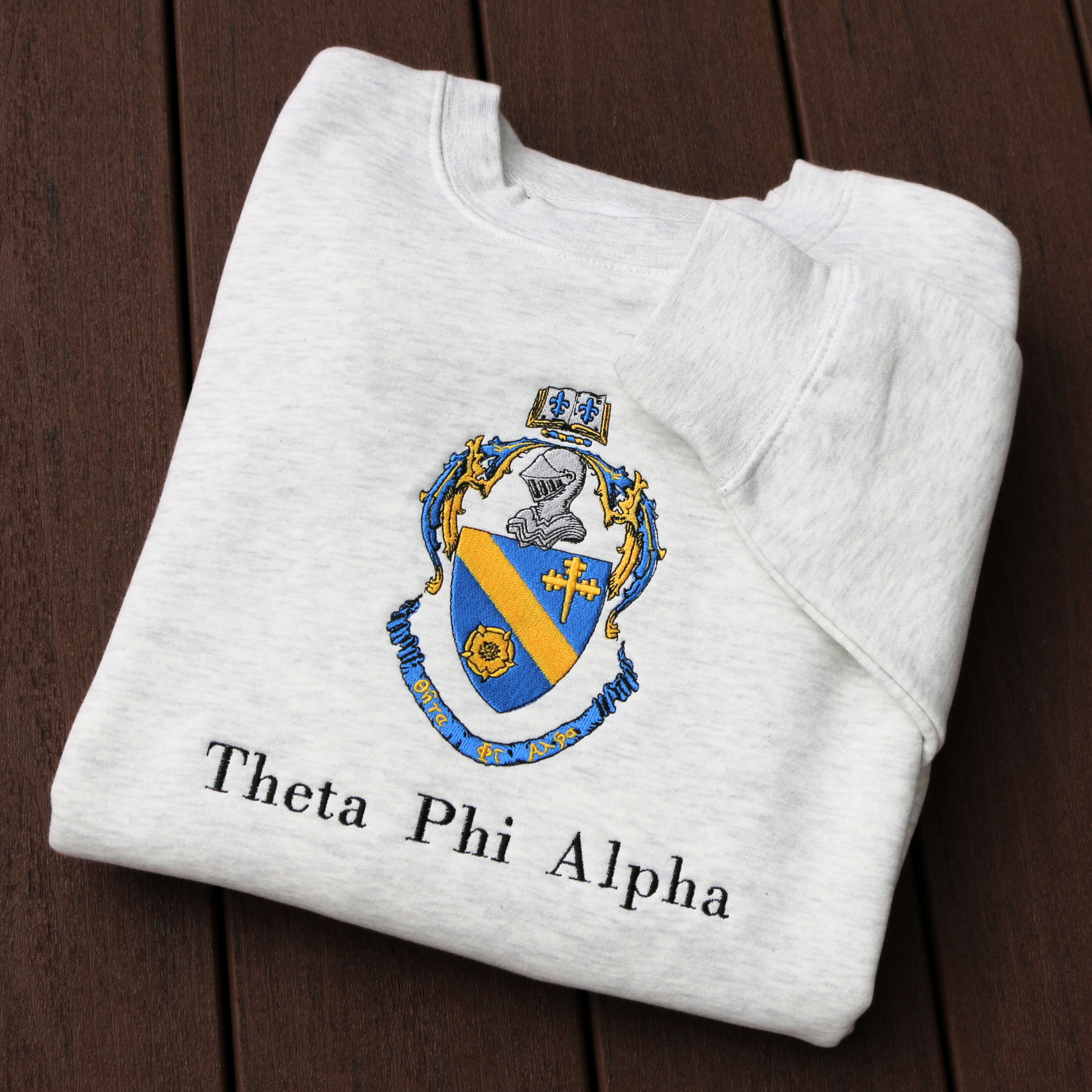Theta Phi Alpha's crest embroidered on a light grey sweatshirt. Featuring a gold and blue crest, with an open book and head of armor atop it. The book is imprinted with two blue fleur-de-lis. The motto is written  on the blue banner at the bottom of the crest. The sorority name is embroidered beneath in black.