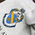 Load image into Gallery viewer, Detailed view of Theta Phi Alpha's crest embroidered on a light grey sweatshirt. Featuring a gold and blue crest, with an open book and head of armor atop it. The book is imprinted with two blue fleur-de-lis. The motto is written  on the blue banner at the bottom of the crest. The sorority name is embroidered beneath in black.
