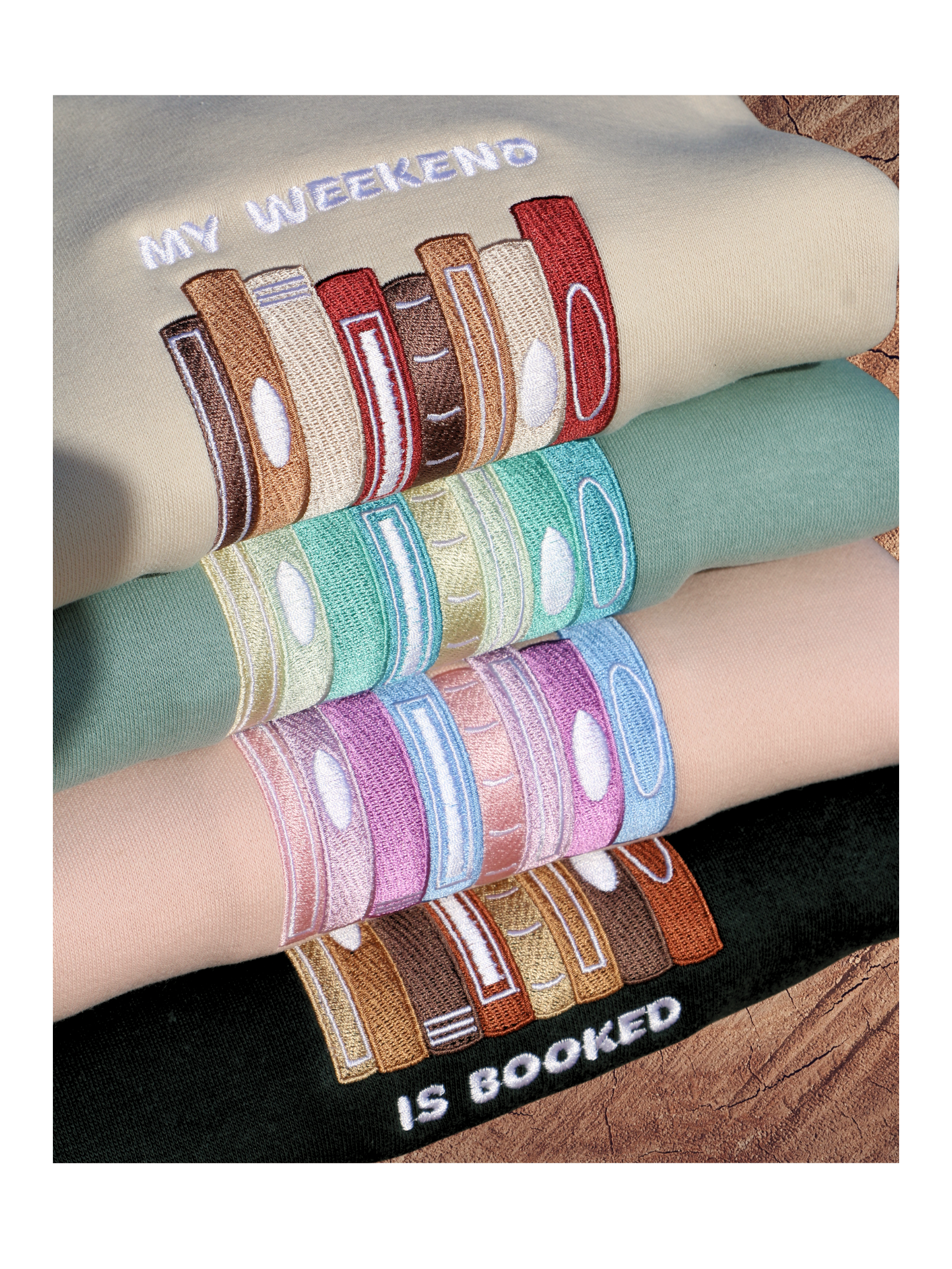 Stack of folded sweatshirts in beige, mint, pink, and dark green, each embroidered with colorful book spines and 'My Weekend is Booked' text.