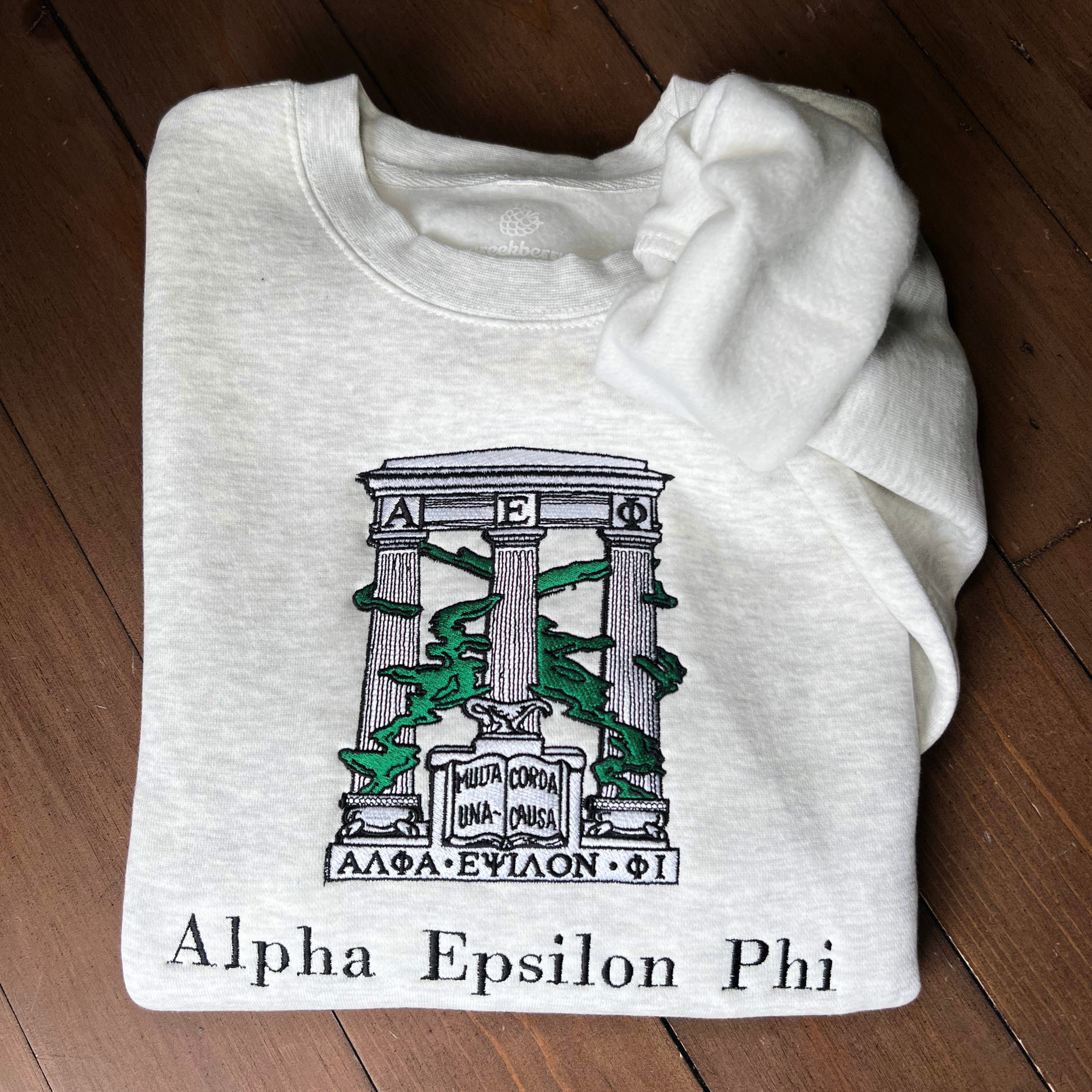 Folded light gray sweatshirt with an embroidered design of Alpha Epsilon Phi's emblem featuring white columns and green foliage. The columns support a structure with the Greek letters 'ΑΕΦ'. Below, the motto 'Multa Corda, Una Causa' is prominently displayed.