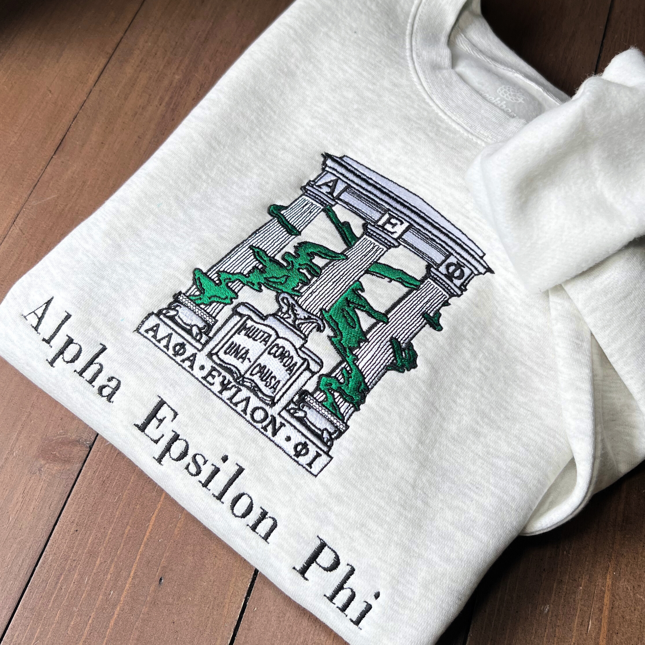 Folded light gray sweatshirt with an embroidered design of Alpha Epsilon Phi's emblem featuring white columns and green foliage. The columns support a structure with the Greek letters 'ΑΕΦ'. Below, the motto 'Multa Corda, Una Causa' is prominently displayed.