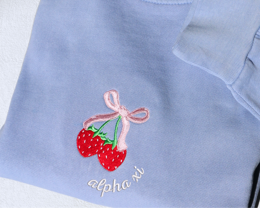 A folded light blue sweatshirt with an embroidered design of strawberries tied with a pink bow and the sorority name embroidered in white underneath.